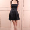 Woman wearing a black satin mini dress with wide straps and a square neckline, posing elegantly against a light pink paneled background.